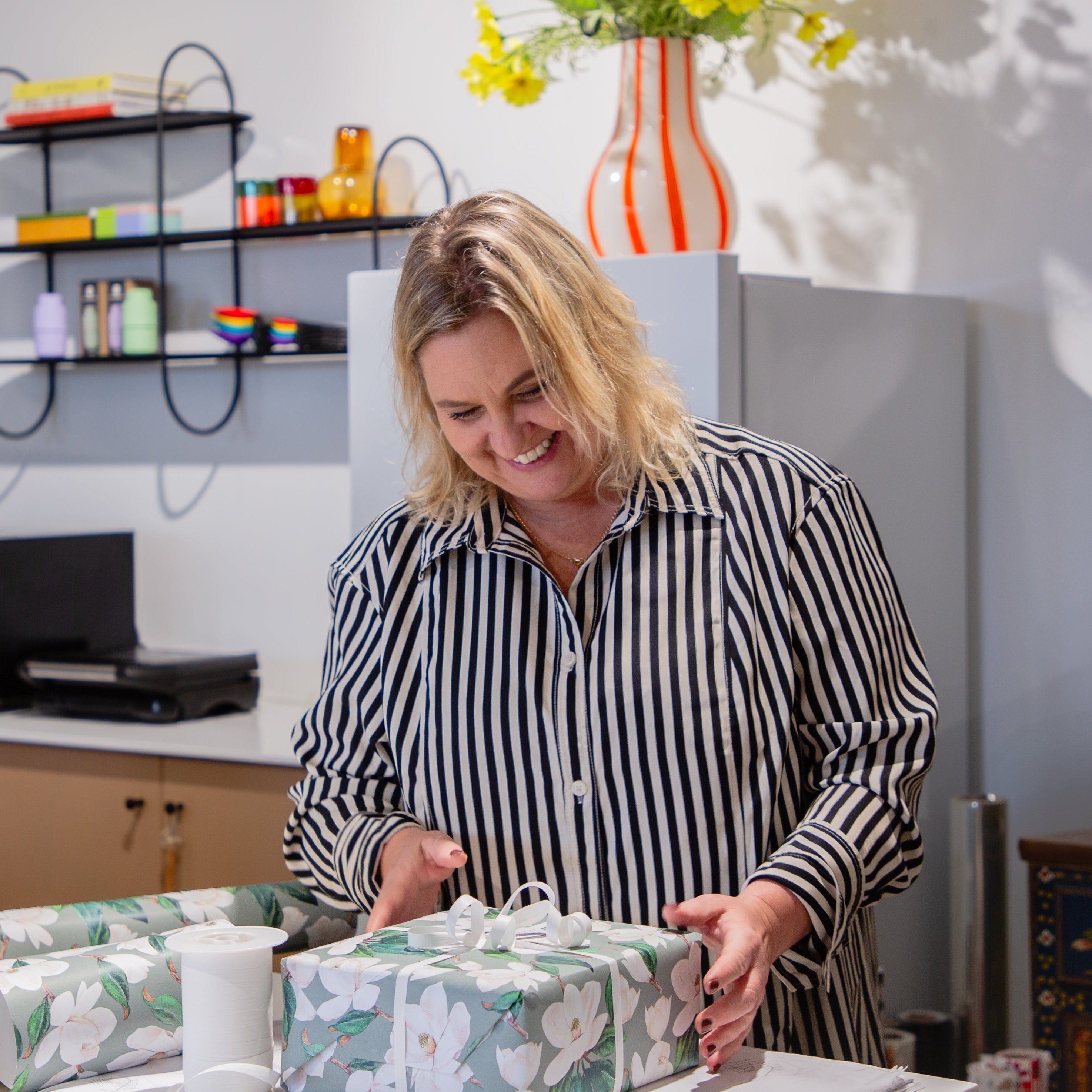 Woman in a kitchen wrapping a gift, with shelves and decor in the background.